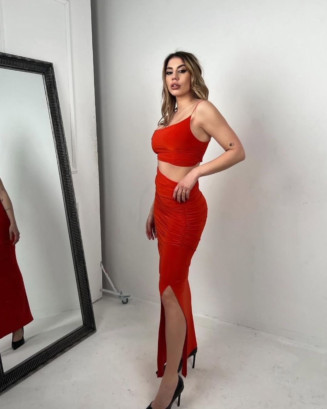 Woman in vibrant red crop top and ruched skirt set with side slit, posing in high heels