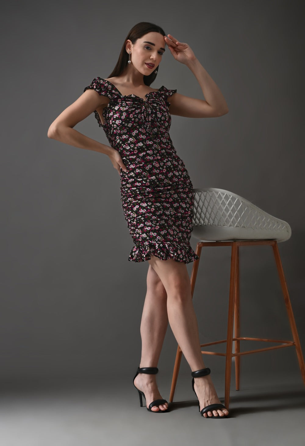 Woman posing in a black floral ruffle mini dress and black heels against a gray background