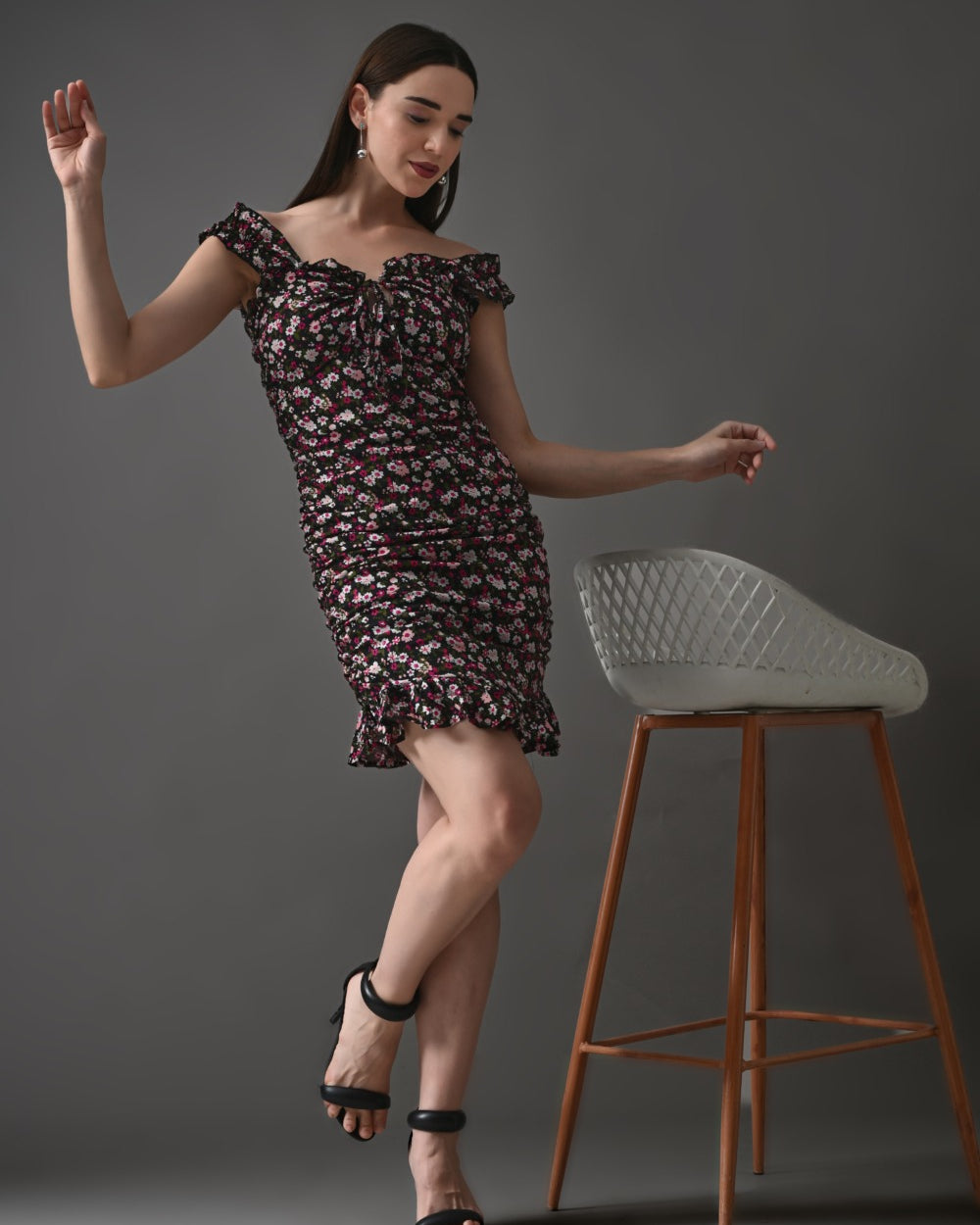 Woman in off-shoulder black floral ruffle mini dress and heels posing by modern chair