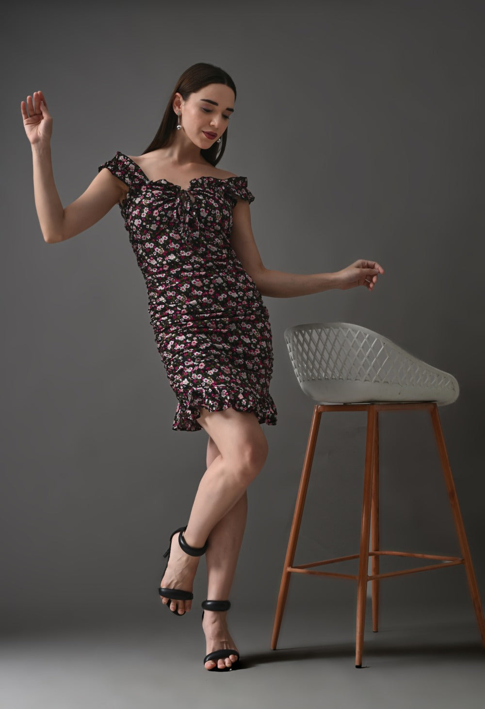 Woman in off-shoulder black floral ruffle mini dress and heels posing by modern chair
